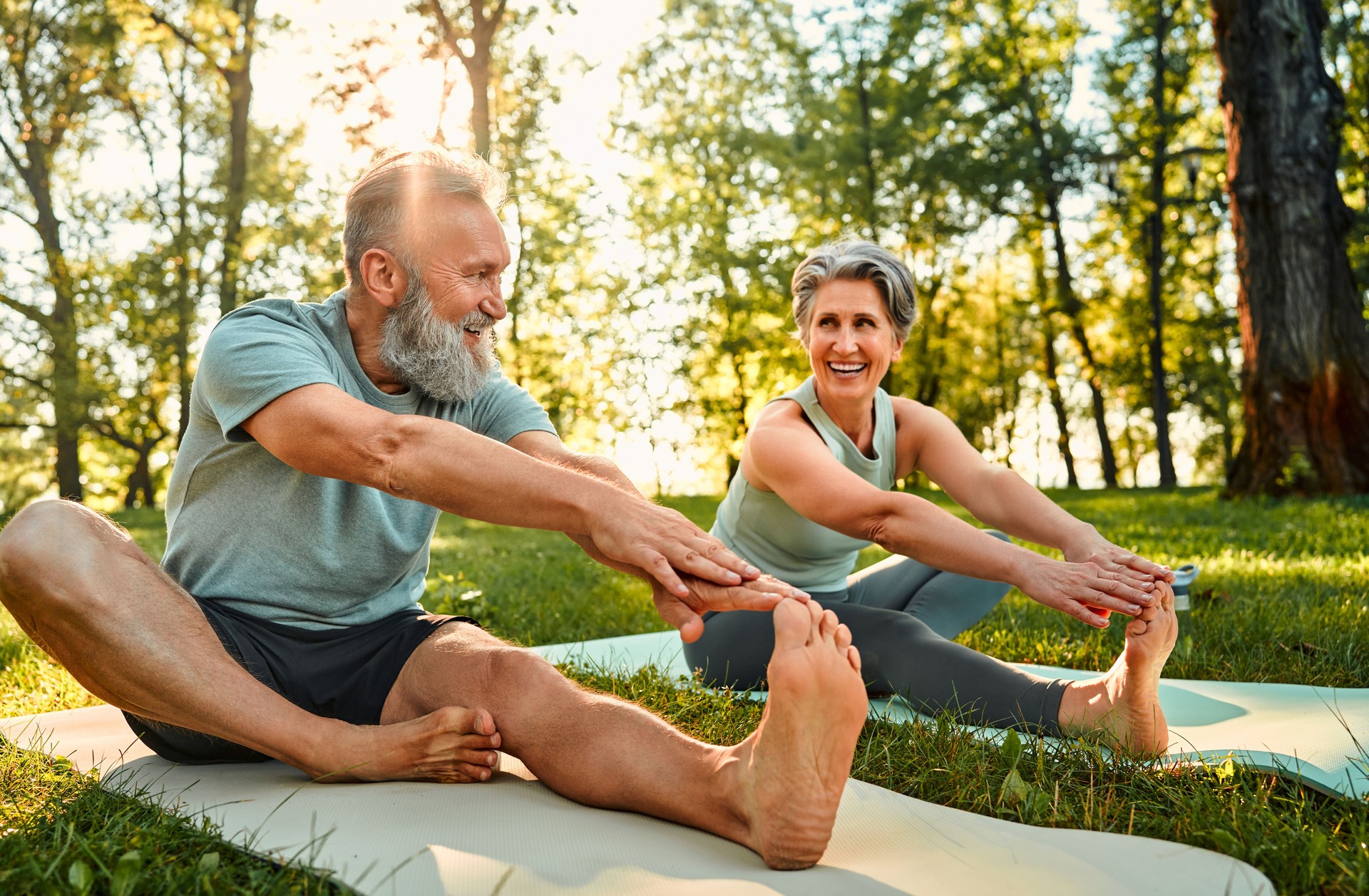 Ejercicios flexibles para el cuerpo. Hombre y mujer deportistas con cabello gris que se estiran en colchonetas de yoga con las manos en una pierna durante el entrenamiento al aire libre. Feliz pareja casada con los pies descalzos calentando juntos en el pa